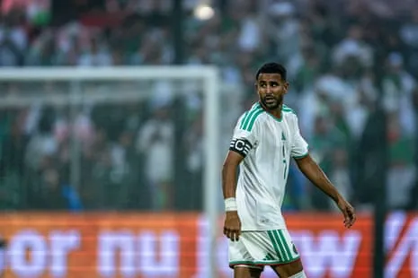 Riyad Mahrez, the Algeria winger and captain, looks over his shoulder while walking to take up a position during a match for his country. He is wearing Algeria’s kit, white with green detailing, and a black captain’s armband.